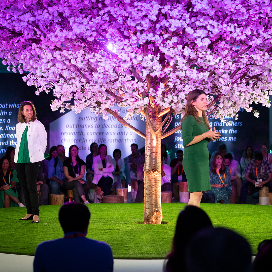 Event setting with a audience surrounding a stage that is dressed as a garden with green grass and a pink blossom tree. Two white female stand on stage talking to the audience.